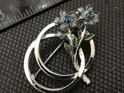 Sale Photo Thumbnail #97: A silver-tone costume jewelry brooch featuring two enameled flowers with blue rhinestone centers set against an intertwined double-ring frame. Includes green painted leaf accents. Features a standard rollover safety clasp on the back. Shows light surface 