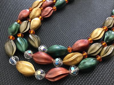 Sale Photo Thumbnail #187: A vintage 3-strand beaded necklace featuring swirl/leaf-molded plastic beads in earth tones (green, gold, bronze, terracotta/coral), accented with faceted crystal and amber spacer beads. Includes decorative metal bar connectors and a hook-and-tail clasp e