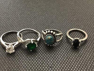 Sale Photo Thumbnail #192: Collection of four silver-toned costume rings featuring various stone designs, including a clear cz-style solitaire with accent stones, a green stone in a bypass-style band, a turquoise-colored cabochon with detailed border, and a black marquise-cut stone