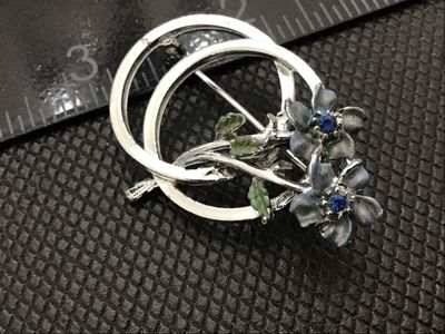 Sale Photo Thumbnail #98: A silver-tone costume jewelry brooch featuring two enameled flowers with blue rhinestone centers set against an intertwined double-ring frame. Includes green painted leaf accents. Features a standard rollover safety clasp on the back. Shows light surface 