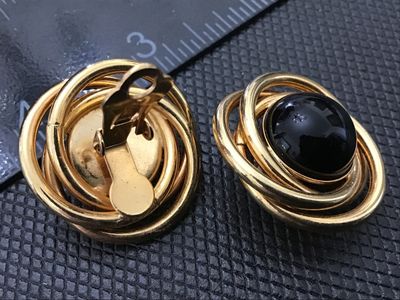 Sale Photo Thumbnail #79: Pair of vintage gold-tone clip-on earrings featuring a multi-ring swirl design centered with a glossy black cabochon stone. The clip backs are functional and include soft comfort pads. Measure approximately 1 inch in diameter. In good condition with minim
