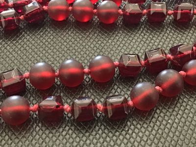 Sale Photo Thumbnail #27: A long single-strand necklace featuring alternating deep red frosted round glass beads and faceted geometric glass beads, hand-knotted on red cord. Finished with a silver-tone lobster clasp closure. Measures approximately 24 to 26 inches in total length. 