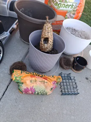 Sale Photo Thumbnail #114: Assorted lawn and garden supply lot including WAGNER'S Songbird Supreme Wild Bird Food, potting mix, multiple plastic and ceramic planters/pots, woven decorative bird roosts/nests, and a suet cage feeder. Items show signs of previous outdoor use and soil 