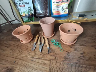 Sale Photo Thumbnail #403: Assorted gardening and plant care supplies including terracotta pots with saucers, hand tools (trowel, pruners, rake), HARRIS Horticultural Charcoal, Organic Perlite, BAYER ADVANCED Home Pest spray, and plant ties.