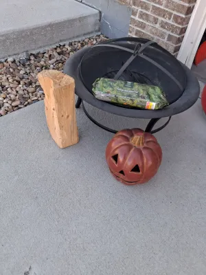 Sale Photo Thumbnail #118: Metal fire pit with mesh spark screen lid and interior grate, American Home by Yankee Candle Fresh Balsam Fir firelog (4.5 lb), ceramic jack-o'-lantern decor, and wood log.