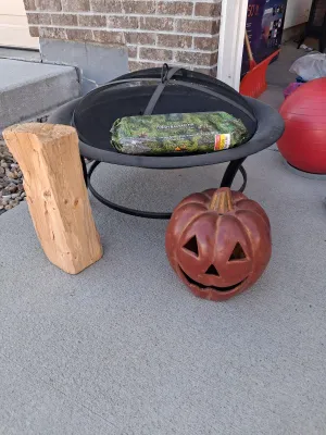 Sale Photo Thumbnail #119: Metal fire pit with mesh spark screen lid and interior grate, American Home by Yankee Candle Fresh Balsam Fir firelog (4.5 lb), ceramic jack-o'-lantern decor, and wood log.
