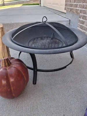 Sale Photo Thumbnail #124: Metal fire pit with mesh spark screen lid and interior grate, American Home by Yankee Candle Fresh Balsam Fir firelog (4.5 lb), ceramic jack-o'-lantern decor, and wood log.