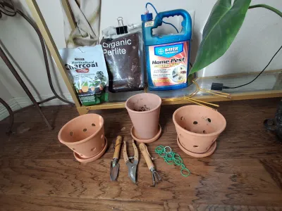 Sale Photo Thumbnail #402: Assorted gardening and plant care supplies including terracotta pots with saucers, hand tools (trowel, pruners, rake), HARRIS Horticultural Charcoal, Organic Perlite, BAYER ADVANCED Home Pest spray, and plant ties.