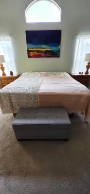 Sale Photo Thumbnail #34: Spacious bedroom suite featuring a king-size bed with lace-accented linens, a gray tufted storage ottoman bench at the foot, and flanking wooden nightstands with lamps.
