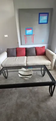 Sale Photo Thumbnail #36: Contemporary living room set featuring a beige upholstered sofa with red and grey throw pillows, a dark metal glass-top coffee table, and a large abstract canvas painting.