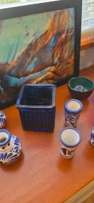 Sale Photo Thumbnail #17: Collection of decorative glassware and ceramics, including blue and white Talavera-style pottery shot glasses, a dark cobalt waffle-textured glass container, and framed artwork on a wooden surface.