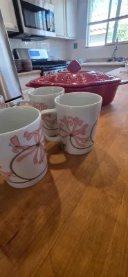 Sale Photo Thumbnail #7: Set of white porcelain mugs with pink floral lily motifs, displayed alongside a deep red embossed ceramic casserole dish on a wooden kitchen island.