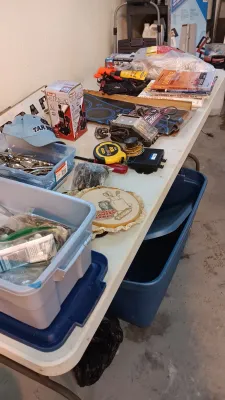 Sale Photo Thumbnail #8: Folding table laden with garage and household items, including a Pro-Lift bottle jack in box, hand tools, automotive manuals, and plastic storage totes.