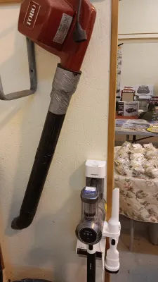 Sale Photo Thumbnail #34: Vintage red Toro electric leaf blower with duct tape repair on the nozzle, mounted next to a modern Tineco cordless vacuum cleaner station.