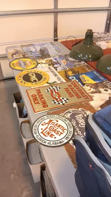 Sale Photo Thumbnail #59: A table arranged with assorted vintage-style metal advertising signs, including Chevrolet, GMC, Atlantic Coast Line, and Ford automotive and railroad memorabilia, alongside green industrial pendant light fixtures.