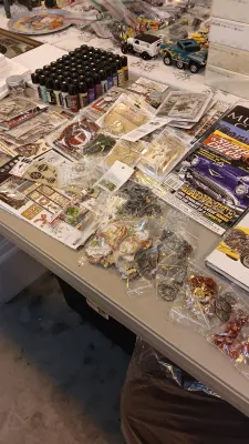 Sale Photo Thumbnail #41: Large assortment of crafting supplies, including acrylic paint bottles, scrapbooking paper and embellishments, jewelry-making charms, and automotive magazines arranged on a folding table.