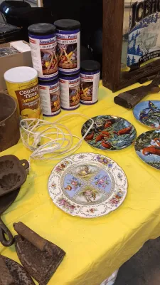 Sale Photo Thumbnail #24: Collection of tabletop items including decorative collector plates, vintage cast iron cookware and irons, cans of Survival Seeds 4 Patriots, and a Country Time Lemonade mix canister.