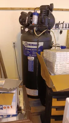 Sale Photo Thumbnail #60: Kobalt 60-gallon vertical cast iron air compressor standing in a workshop setting, accompanied by a stack of boxed air filters.