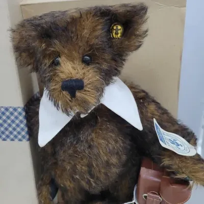 Vintage dark brown mohair teddy bear featuring a white collar, miniature brown leather bag, and original button and tag in ear.