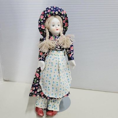 Vintage porcelain doll with yarn pigtails, dressed in a floral pattern dress, bonnet, apron, and red clogs.