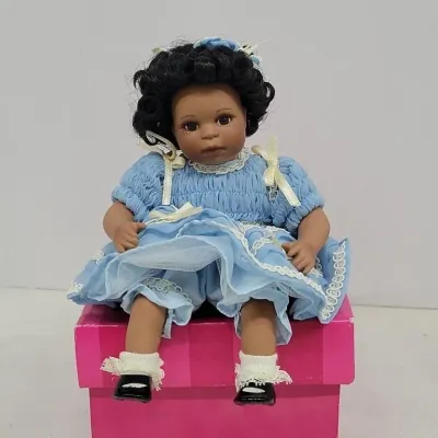 Collectible porcelain African American child doll featuring short curly black hair, wearing a light blue smocked dress with white trim,...