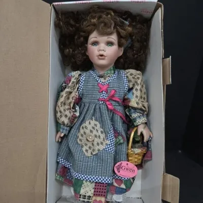 Vintage porcelain doll named Gwen, featuring curly brown hair, green eyes, and a patterned patchwork dress, presented in its original box.