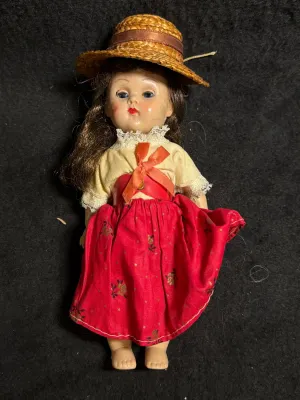 Vintage hard plastic storybook doll wearing a straw hat, lace-trimmed top, and red floral skirt, circa mid-20th century.