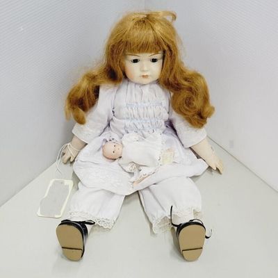 Porcelain doll with reddish-brown hair dressed in a white lace trim outfit, holding a miniature baby doll.