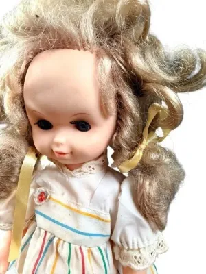 Vintage vinyl doll with blonde curly pigtails tied with yellow ribbons, wearing a striped white dress with eyelet lace trim.