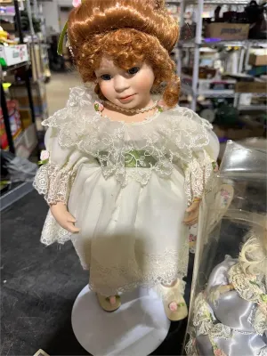 Vintage porcelain collector doll featuring reddish-brown curled hair, blue eyes, and an ornate lace-trimmed white dress with floral accents.