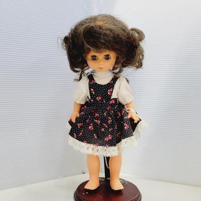 Vintage plastic girl doll with brown curly hair and large eyes, wearing a black floral pinafore dress over a white lace-trimmed blouse,...