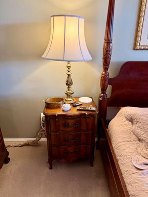French Provincial style wooden three-drawer nightstand topped with a polished brass table lamp and decorative accessories, positioned...