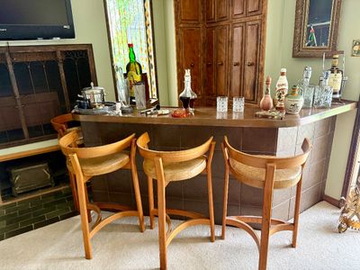Mid-century modern home bar set featuring curved wooden bar stools with tan vinyl seats alongside various vintage glassware, decanters,...