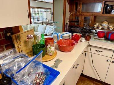 Kitchen counter filled with various glassware, ceramic cabbage-leaf serving bowl, red teapots, stackable dishware, and bagged silver...