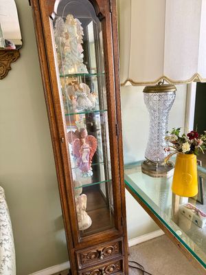 Tall narrow wooden curio cabinet housing various angel figurines alongside a vintage cut-glass table lamp on a glass console table.