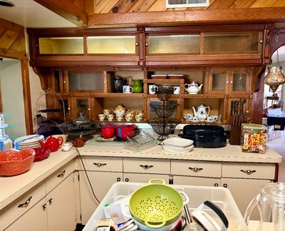 Large assortment of kitchenware and tableware displayed on counter space and built-in wooden shelving. Items include tea sets, roasting...