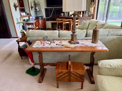 Wooden trestle sofa table with lace runner, holding decor items including a brass-toned table lamp, flanked by a vintage wooden accordion...