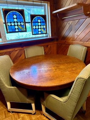 Round solid wood dining table set with four upholstered rolling armchair seats, complemented by hanging stained glass window panels.