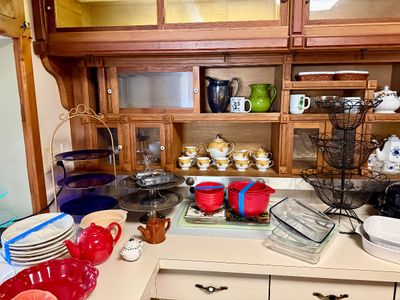 Kitchen cabinet counter filled with various tableware items including teapots, teacups, nesting bowls, a black wire tier basket, glass...