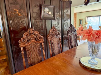 Set of ornate carved wood dining chairs with cane backs, shown alongside a wooden dining table and an intricately carved dark wood screen.