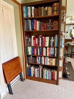 Tall wood bookcase filled with various vintage hardcovers, paperbacks, and antique leather-bound books, alongside a folding wood TV tray...