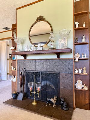 Tiled fireplace hearth flanked by wooden shelving units, featuring a round ornate mirror, glass compotes, oil lamps, angel figurines, and...