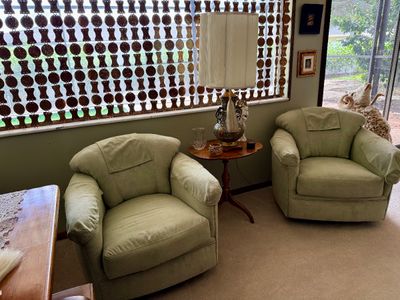 Pair of sage green suede swivel armchairs flank a vintage wooden side table topped with a tall lamp with fringe details.