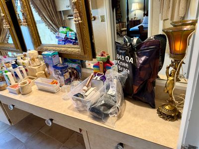 Bathroom vanity counter filled with various personal care products, hair styling tools, medical knee braces, toiletries, and an ornate...