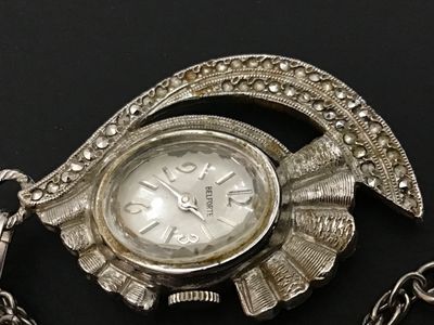Sale Photo Thumbnail #32: Vintage Belforte pendant watch styled as a necklace. Features an ornate silver-tone, fan-shaped housing set with marcasite-style accents, an oval watch face with silvertone hands and numbers, and a matching chain. The back of the watch casing is marked 'B