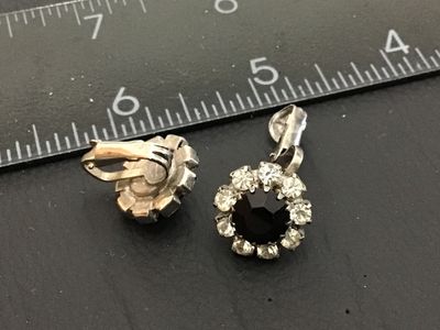 Sale Photo Thumbnail #241: Pair of vintage silver-tone clip-on earrings featuring a central faceted black rhinestone surrounded by a halo of clear rhinestones. Each earring measures approximately 0.5 inches in diameter. Show minor age-appropriate surface wear to metal backing.