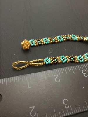 Sale Photo Thumbnail #127: Handmade woven seed bead bracelet featuring a turquoise-blue and gold-tone floral daisy chain design. Includes a beaded loop and beaded ball toggle closure. Measures approximately 7 inches long. In good condition with no missing beads visible.