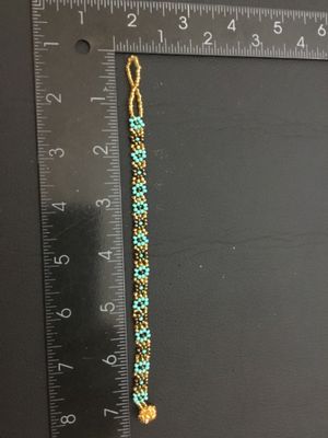 Sale Photo Thumbnail #121: Handmade woven seed bead bracelet featuring a turquoise-blue and gold-tone floral daisy chain design. Includes a beaded loop and beaded ball toggle closure. Measures approximately 7 inches long. In good condition with no missing beads visible.