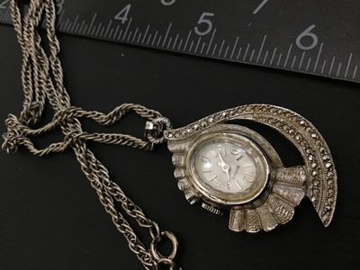 Sale Photo Thumbnail #36: Vintage Belforte pendant watch styled as a necklace. Features an ornate silver-tone, fan-shaped housing set with marcasite-style accents, an oval watch face with silvertone hands and numbers, and a matching chain. The back of the watch casing is marked 'B