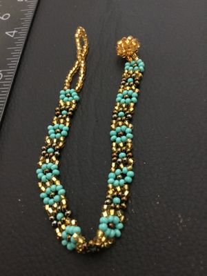 Sale Photo Thumbnail #128: Handmade woven seed bead bracelet featuring a turquoise-blue and gold-tone floral daisy chain design. Includes a beaded loop and beaded ball toggle closure. Measures approximately 7 inches long. In good condition with no missing beads visible.